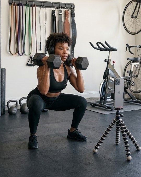 Woman doing a dumbbell squat at home with her phone on a tripod.